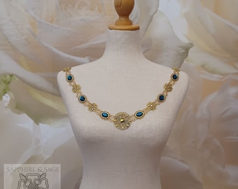 May include: A gold necklace with a large, ornate, circular pendant and smaller oval-shaped stones. The stones are a deep teal blue color. The necklace is on a white mannequin.
