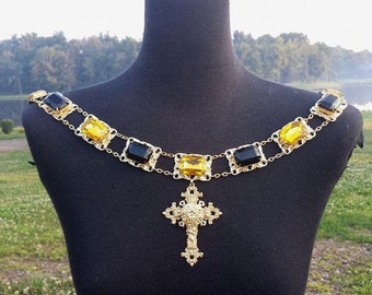 May include: A gold-toned necklace with black and yellow rectangular gemstones, connected by delicate chain links. A detailed cross pendant hangs from the center. The necklace is displayed on a black mannequin.