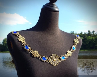 May include: A gold chain necklace with a large circular pendant and oval blue gemstones. The necklace is on a black mannequin.