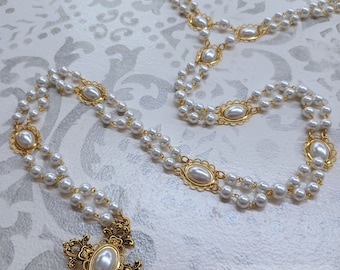 May include: A double strand pearl necklace with a gold cross pendant. The cross has a large pearl in the center and intricate detailing. The necklace is on a white background with a floral pattern.