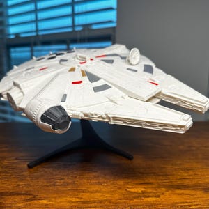 Può includere: Un modello dettagliato dell'astronave Millennium Falcon, prevalentemente bianco con accenti grigi e rossi. Il modello è esposto su un supporto nero, mostrando il suo design iconico. I dettagli intricati della nave sono visibili.
