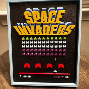 Op de afbeelding: Een ingelijst kunstwerk met de tekst "SPACE INVADERS" in geel, blauw en wit. Het kunstwerk toont gepixelde alien-indringers in groen en paars, met rode en witte spelelementen aan de onderkant. De lijst is lichtblauw.