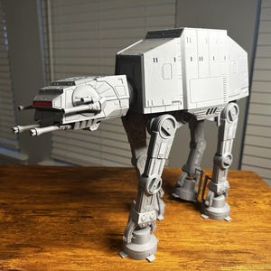 May include: A gray AT-AT model from Star Wars, standing on a wooden surface. The model features detailed mechanical elements, including cannons, leg joints, and a rectangular body. The model is a light gray color.
