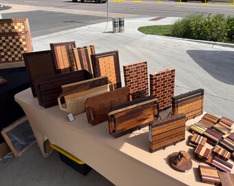 Könnte beinhalten: Ausstellung handgefertigter Holzartikel, darunter Schneidebretter, Serviertabletts und dekorative Blöcke. Die Artikel weisen verschiedene Holzmaserungen und Muster auf, mit einer Mischung aus rechteckigen und quadratischen Formen. Einige haben Griffe oder dekorative Einlegearbeiten.