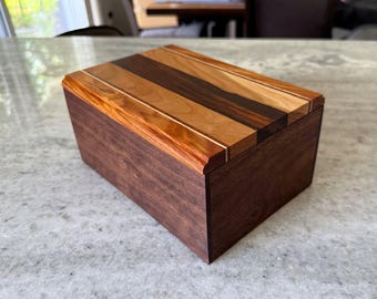 May include: A rectangular wooden box featuring a dark brown walnut base and a lid constructed from alternating strips of light cherry, medium-toned wood, and dark walnut. The lid has a beveled edge and a smooth, polished finish throughout the piece.
