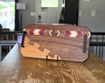 May include: A rectangular wooden serving tray featuring a dark walnut base with a lighter cherry wood inlay in a puzzle-piece pattern. The top edge displays a decorative chevron inlay of purple and light wood. Two black metal handles are attached.