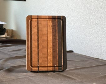 Könnte beinhalten: Ein rechteckiges Holzschneidebrett mit Saftrille. Das Brett besteht aus verschiedenen Holztönen, darunter hellbraune, mittelbraune und dunkelbraune Streifen. Es wird auf einem schwarzen Metallständer präsentiert.