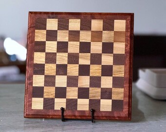 May include: A handcrafted wooden chess board with a dark brown frame and a checkerboard pattern of light and dark squares. The board is displayed on a black metal stand, showcasing the contrasting wood tones and craftsmanship.