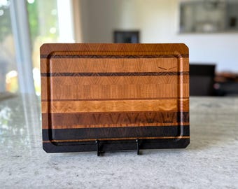 May include: A rectangular wooden cutting board featuring horizontal stripes of varying wood tones ranging from light tan to dark brown and black. The board has a smooth surface with a recessed groove along the perimeter and is displayed on a black stand.