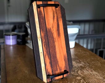 May include: A rectangular wooden serving tray featuring a striped pattern of dark brown and light orange wood grain. The tray includes two black metal handles attached to the top and bottom edges, providing a functional grip for carrying.