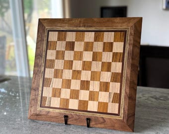 May include: A square wooden chessboard featuring a checkered pattern of alternating light maple and medium brown walnut wood squares. The board is framed by a wide, dark walnut border with a mitered edge and rests on a black metal display stand.