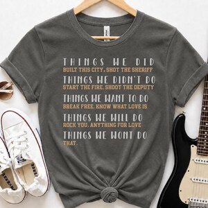 May include: A dark gray short-sleeve t-shirt featuring white and tan text that lists various song titles and phrases including Things We Did, Built This City, Shot The Sheriff, Things We Didn't Do, Start The Fire, Shoot The Deputy, and more.