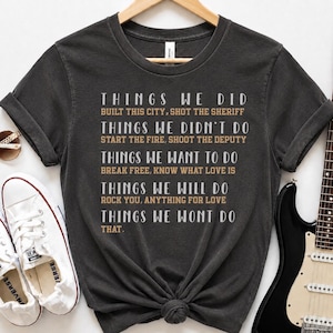 May include: A charcoal gray short-sleeve t-shirt featuring white and tan text that reads: THINGS WE DID BUILT THIS CITY, SHOT THE SHERIFF; THINGS WE DIDN'T DO START THE FIRE, SHOOT THE DEPUTY; THINGS WE WANT TO DO BREAK FREE, KNOW WHAT LOVE IS.