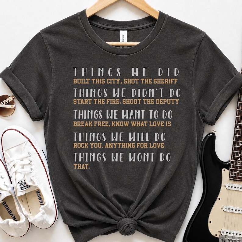 May include: A charcoal gray short-sleeve t-shirt featuring white and tan text that reads: THINGS WE DID BUILT THIS CITY, SHOT THE SHERIFF; THINGS WE DIDN'T DO START THE FIRE, SHOOT THE DEPUTY; THINGS WE WANT TO DO BREAK FREE, KNOW WHAT LOVE IS.