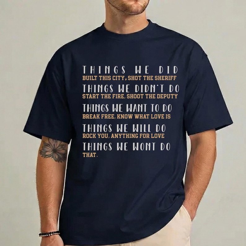 May include: A navy blue short-sleeved t-shirt featuring white and tan text that reads: THINGS WE DID BUILT THIS CITY, SHOT THE SHERIFF; THINGS WE DIDN'T DO START THE FIRE, SHOOT THE DEPUTY; THINGS WE WANT TO DO BREAK FREE, KNOW WHAT LOVE IS; THINGS WE WILL DO ROCK YOU, ANYTHING FOR LOVE; THINGS WE WONT DO THAT.