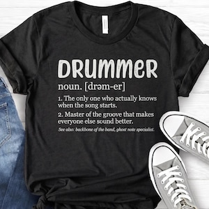 Op de afbeelding: Zwart T-shirt met het woord "DRUMMER" in witte letters, met een definitie eronder. De definitie bevat de uitspraak en twee uitspraken over drummers. Het shirt wordt gecombineerd met grijze sneakers.