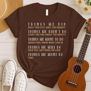 May include: A brown short-sleeved t-shirt featuring white and tan text that lists various actions under headings like Things We Did, Things We Didn't Do, Things We Want To Do, Things We Will Do, and Things We Wont Do That, including song titles.
