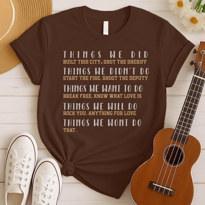 May include: A brown short-sleeved t-shirt featuring white and tan text that lists various actions under headings like Things We Did, Things We Didn't Do, Things We Want To Do, Things We Will Do, and Things We Wont Do That, including song titles.