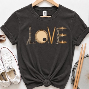 Puede incluir: Una camiseta gris carbón de manga corta con un diseño gráfico donde la palabra LOVE está formada por piezas de batería. La L es un soporte, la O es una caja, la V son dos baquetas y la E es una pila de tres platillos.