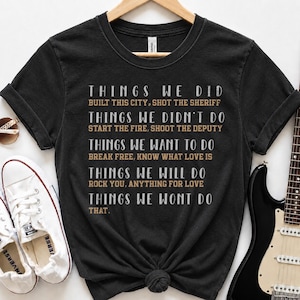 May include: A black short-sleeved t-shirt featuring white and tan text that reads: THINGS WE DID BUILT THIS CITY, SHOT THE SHERIFF; THINGS WE DIDN'T DO START THE FIRE, SHOOT THE DEPUTY; THINGS WE WANT TO DO BREAK FREE, KNOW WHAT LOVE IS; THINGS WE WILL DO ROCK YOU, ANYTHING FOR LOVE; THINGS WE WONT DO THAT.