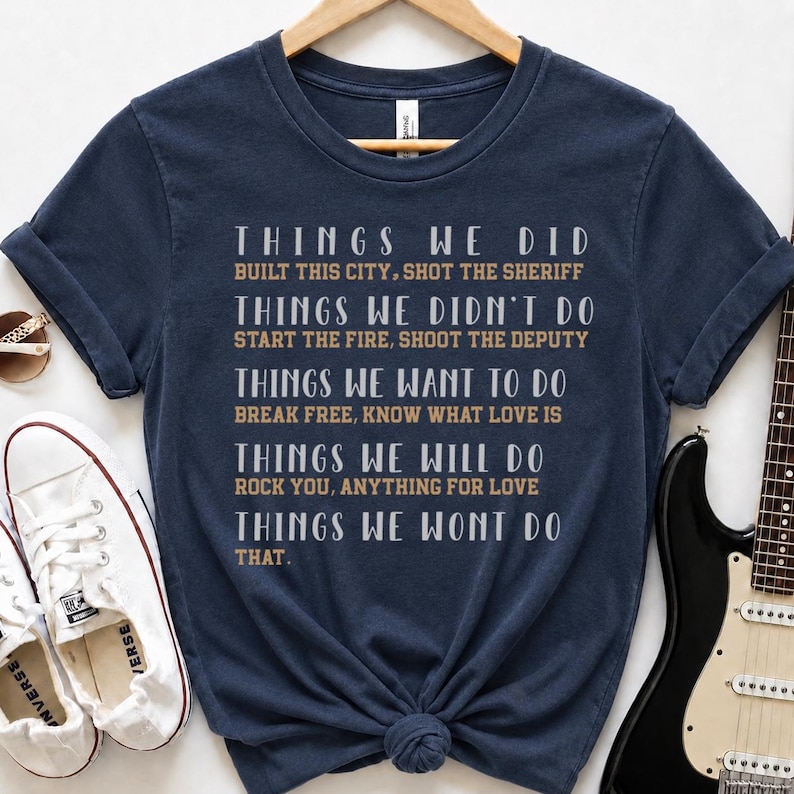 May include: A navy blue short-sleeved t-shirt featuring white and tan text that lists various song titles and phrases including Things We Did, Built This City, Shot The Sheriff, Things We Didn't Do, Start The Fire, Shoot The Deputy, and more.