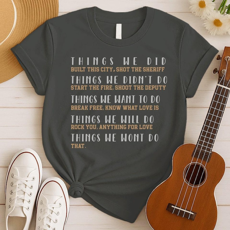 May include: A dark gray short-sleeve t-shirt featuring white and tan text that reads: THINGS WE DID BUILT THIS CITY, SHOT THE SHERIFF; THINGS WE DIDN'T DO START THE FIRE, SHOOT THE DEPUTY; THINGS WE WANT TO DO BREAK FREE, KNOW WHAT LOVE IS; THINGS WE WILL DO ROCK YOU, ANYTHING FOR LOVE; THINGS WE WONT DO THAT.