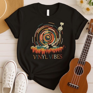 May include: Black t-shirt featuring a retro-style graphic of a record player with a colorful vinyl record design. The text "VINYL VIBES" is printed below the graphic in a vintage font. A pair of white sneakers and a ukulele are in the image.