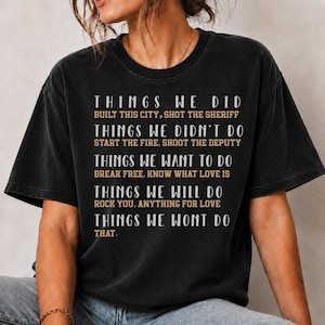 May include: A black short-sleeve t-shirt featuring white and tan text that reads: THINGS WE DID BUILT THIS CITY, SHOT THE SHERIFF; THINGS WE DIDN'T DO START THE FIRE, SHOOT THE DEPUTY; THINGS WE WANT TO DO BREAK FREE, KNOW WHAT LOVE IS; THINGS WE WILL DO ROCK YOU, ANYTHING FOR LOVE; THINGS WE WONT DO THAT.