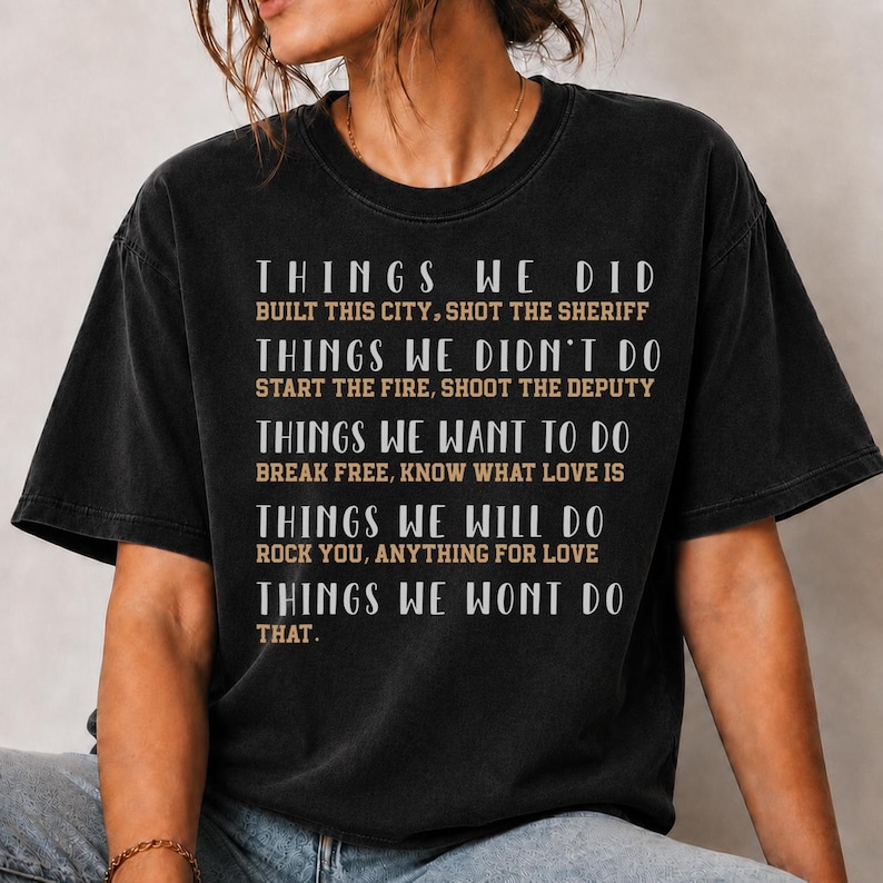 May include: A black short-sleeve t-shirt featuring white and tan text that reads: THINGS WE DID BUILT THIS CITY, SHOT THE SHERIFF; THINGS WE DIDN'T DO START THE FIRE, SHOOT THE DEPUTY; THINGS WE WANT TO DO BREAK FREE, KNOW WHAT LOVE IS; THINGS WE WILL DO ROCK YOU, ANYTHING FOR LOVE; THINGS WE WONT DO THAT.