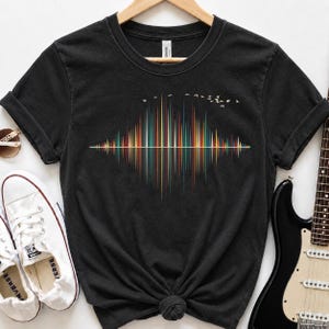 Puede incluir: Una camiseta negra de manga corta con un gráfico central de un patrón de onda sonora colorido en tonos rojo, naranja, amarillo, verde y azul. Sobre la onda sonora, una pequeña bandada de pájaros blancos vuela hacia el lado derecho.