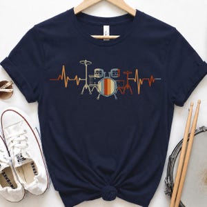 May include: Navy blue t-shirt featuring a retro-style drum set graphic with a heartbeat design. The drum set is in shades of orange, yellow, and blue. The shirt is made of soft material and is suitable for music lovers.