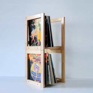 Puede incluir: Un mueble de almacenamiento de discos de vinilo de madera clara de dos niveles. La unidad contiene discos de vinilo, con portadas de álbumes visibles. El diseño es simple y moderno, con un enfoque en la funcionalidad y la exhibición. La unidad es de madera natural.