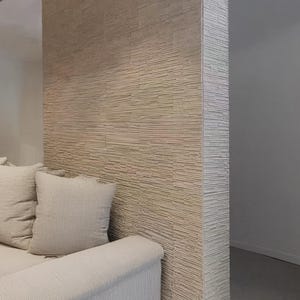 K&ouml;nnte beinhalten: Eine Raumteilerwand mit einer strukturierten Fassade aus gestapeltem Stein in hellem Beige. Die Wand hat eine horizontale, geschichtete Optik mit einer rauen, nat&uuml;rlichen Oberfl&auml;che. Ein hellbeiges Sofa mit passenden Kissen steht vor der Wand.