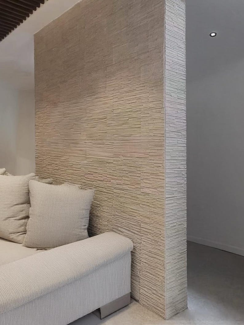 K&ouml;nnte beinhalten: Eine Raumteilerwand mit einer strukturierten Fassade aus gestapeltem Stein in hellem Beige. Die Wand hat eine horizontale, geschichtete Optik mit einer rauen, nat&uuml;rlichen Oberfl&auml;che. Ein hellbeiges Sofa mit passenden Kissen steht vor der Wand.