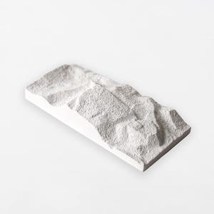 Può includere: Un elemento decorativo rettangolare bianco con un design strutturato a forma di montagna. La superficie ha un aspetto ruvido e granuloso, e i bordi sono puliti e definiti. L'oggetto è posto su uno sfondo bianco.