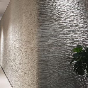 K&ouml;nnte beinhalten: Eine geschwungene Wand mit einer hellgrauen, stark strukturierten Steinverkleidung mit horizontalen, geschichteten Rippen. Die Oberfl&auml;che zeigt ein raues, gestapeltes Aussehen mit tiefen Rillen und Schatten, beleuchtet durch seitliches Licht.