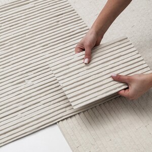 K&ouml;nnte beinhalten: Eine rechteckige, cremewei&szlig;e Steinfliese mit einer linearen, gerippten Textur und parallelen horizontalen Erhebungen. Die Oberfl&auml;che weist ein mattes, nat&uuml;rliches Finish mit subtilen Variationen in der Steinstruktur auf.