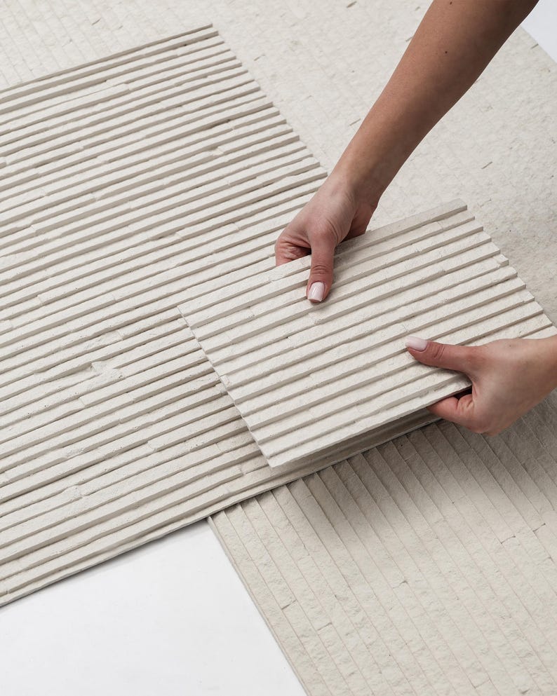 K&ouml;nnte beinhalten: Eine rechteckige, cremewei&szlig;e Steinfliese mit einer linearen, gerippten Textur und parallelen horizontalen Erhebungen. Die Oberfl&auml;che weist ein mattes, nat&uuml;rliches Finish mit subtilen Variationen in der Steinstruktur auf.