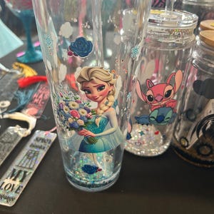 Puede incluir: Vaso de plástico transparente con una imagen de dibujos animados de Elsa de Frozen sosteniendo flores. El vaso tiene una pajita y está lleno de purpurina y pequeñas decoraciones azules en forma de flor. Se ven otros vasos con diferentes diseños.