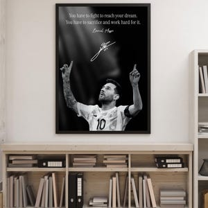 May include: Black and white framed print featuring Lionel Messi, wearing a soccer jersey with the number 10. The print includes a quote and Messi's signature. The image is displayed on a wooden shelf with books and binders.