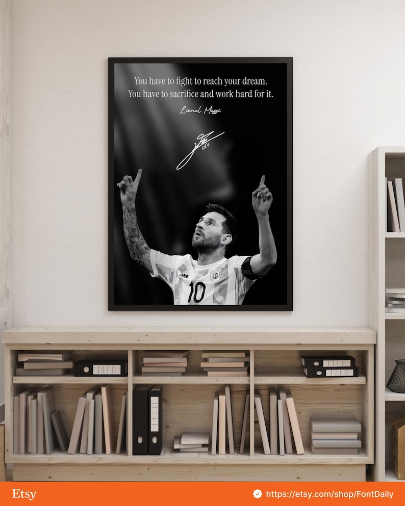 May include: Black and white framed print featuring Lionel Messi, wearing a soccer jersey with the number 10. The print includes a quote and Messi's signature. The image is displayed on a wooden shelf with books and binders.