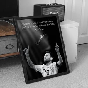 May include: Black and white framed print featuring Lionel Messi. The image shows Messi in a soccer uniform with the number 10, arms raised. The print includes a quote by Messi: "You have to fight to reach your dream."