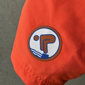 Könnte beinhalten: Orangefarbene Badehose mit einem braunen und blauen Aufnäher, der einen stilisierten Taucher zeigt. Der Aufnäher hat einen weißen Hintergrund und den Text "P" in Blau.