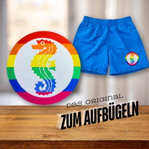 Könnte beinhalten: Ein regenbogenfarbener Bügelaufkleber mit einem weißen Kreis, in dem sich ein Seepferdchen befindet. Der Text "Das Original zum Aufbügeln" ist unter dem Aufkleber sichtbar.