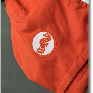 Könnte beinhalten: Orangefarbene Badehose mit einem weißen Kreislogo, das ein Seepferdchen zeigt.