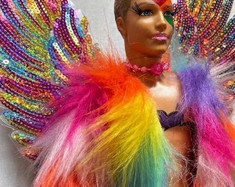 May include: A doll with rainbow colored fur and sequined wings. The doll has a painted face with colorful makeup and is wearing a black bra and a necklace with pink beads.