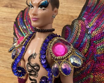 May include: A doll with black hair, blue eyeshadow, and a pink and purple sequined costume. The doll is wearing a blue and gold sequined jacket with a snake tattoo on the chest. The doll is also wearing a pink and purple sequined skirt and wings.