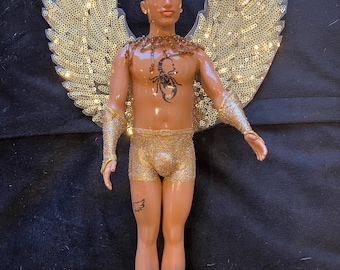 May include: A doll with brown skin, short black hair, and gold glitter wings. The doll is wearing a gold glitter swimsuit and has a scorpion tattoo on its chest.