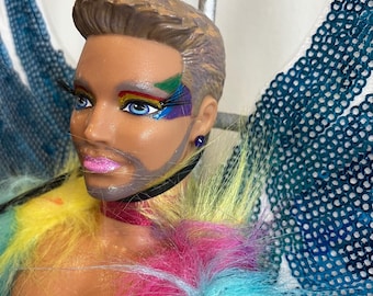 May include: A doll with a rainbow-colored fur coat, a black choker, and a heart tattoo on its chest. The doll has a beard, gray hair, and blue eyes with rainbow makeup. The doll is wearing a blue and teal sequined angel wing.