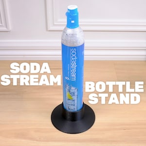 以下が含まれることがあります： 白いキャップが付いた青いSodastream CO2シリンダーが、黒いボトルスタンドに収められています。シリンダーにはSodastreamのロゴと飲み物のグラフィックがあります。「SODA STREAM」と「BOTTLE STAND」の文字は白です。